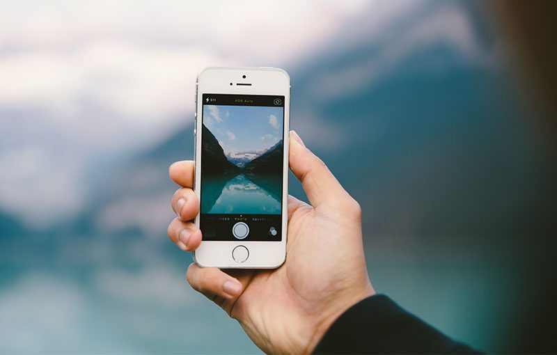 A hand holds a smartphone, capturing a photo of a serene landscape featuring a mountain lake surrounded by cliffs and a cloudy sky. The phone's screen displays the same scene, focusing on the lake and mountains.