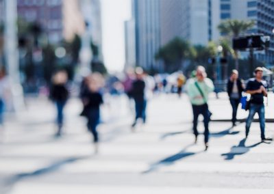 A blurred image of people walking and standing at a crosswalk in an urban setting with tall buildings and palm trees in the background. The focus is soft, giving a sense of motion and activity in the city.