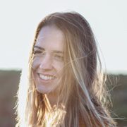A woman with long, light brown hair smiles while standing outdoors. Sunlight illuminates her hair, and a blurred natural landscape can be seen in the background.