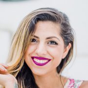 A woman with long, partially blonde hair is smiling. She is wearing vibrant pink lipstick and a floral-patterned top. The background is blurred, focusing attention on her bright and cheerful expression.