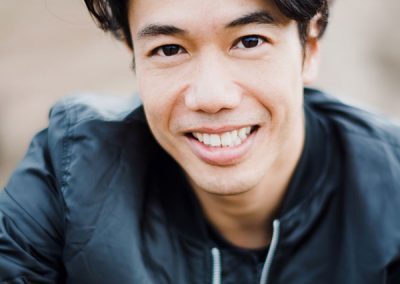 A person with short, dark hair and a radiant smile looks directly at the camera. They are wearing a dark, casual jacket. The background is blurred, emphasizing the subject's face.