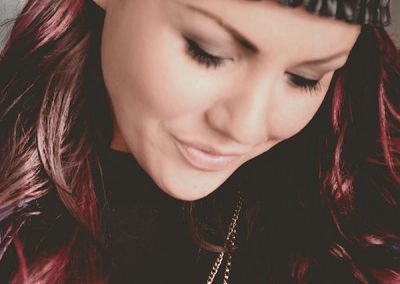 A woman with long, wavy dark hair featuring purple highlights looks downward with a gentle smile. She is wearing a black beanie and a necklace with a pendant. The background is softly blurred, focusing on her serene expression.