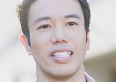 A person with short, dark hair is smiling slightly. They are wearing a light purple button-up shirt. The background is softly blurred, and the lighting is bright and natural, highlighting the person’s face.