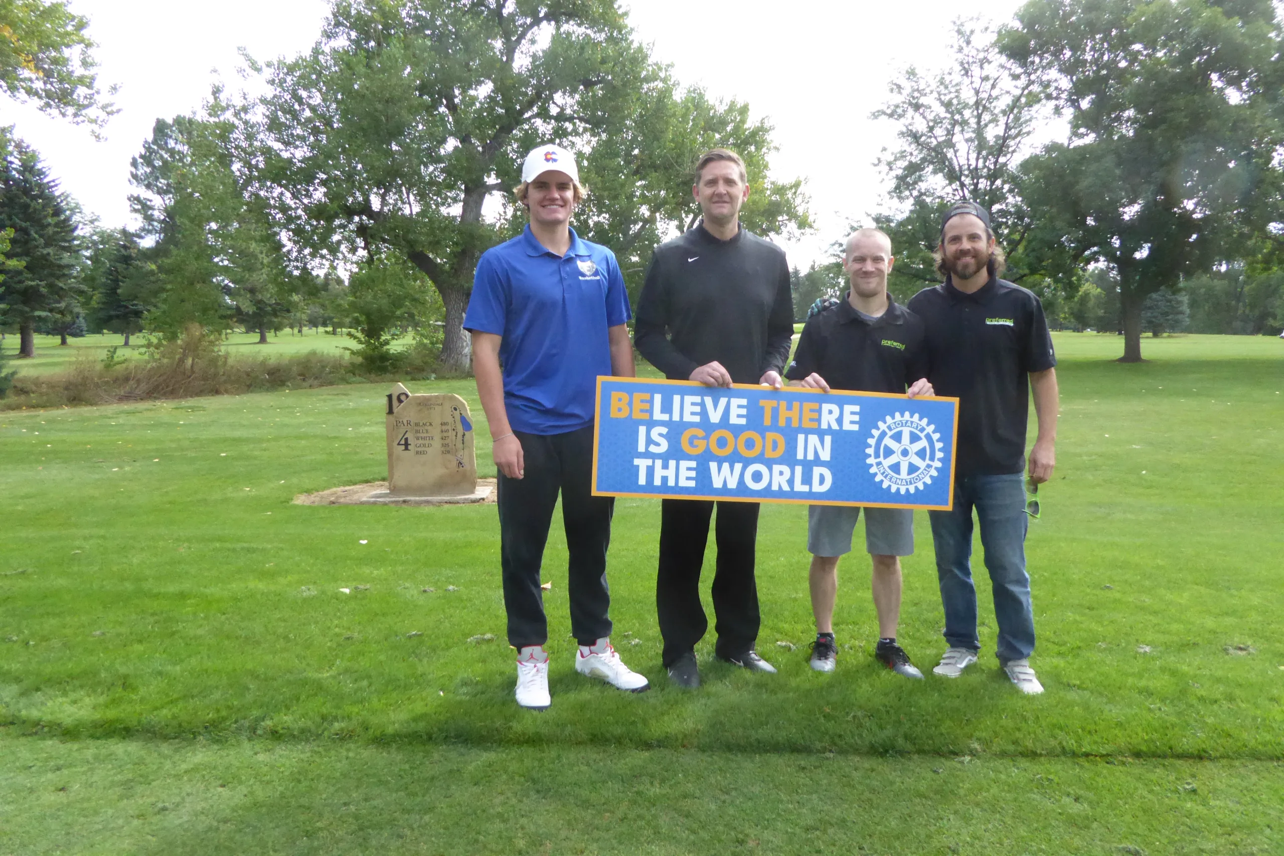 Four men stand on a golf course holding a sign that reads, "BELIEVE THERE IS GOOD IN THE WORLD." One man is in a blue shirt, two in black shirts, and one in a grey shirt. Trees and a grassy field are visible in the background.